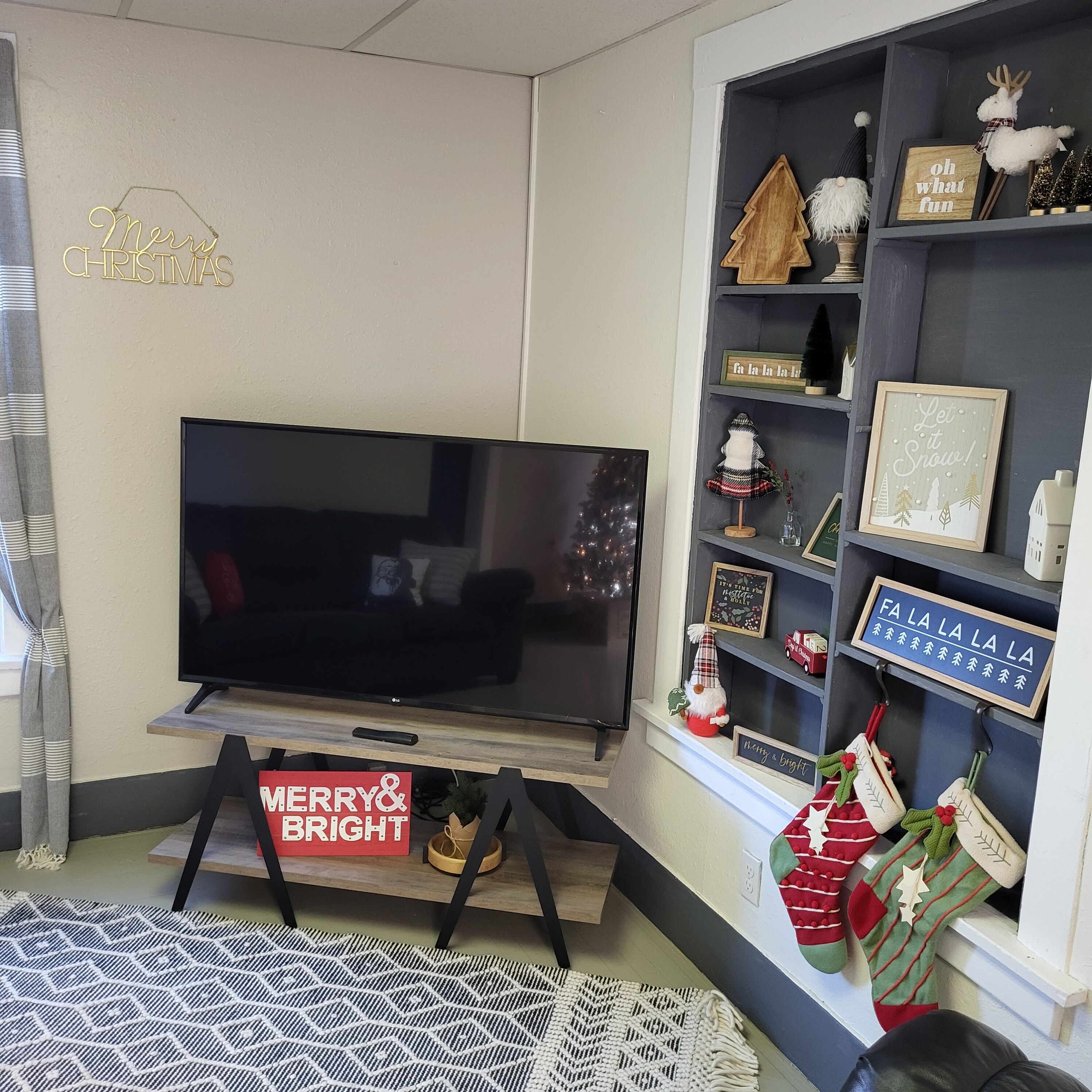 A living room with a flat-screen TV on a stand, a 'Merry & Bright' sign, Christmas decorations, and a bookshelf with various items.