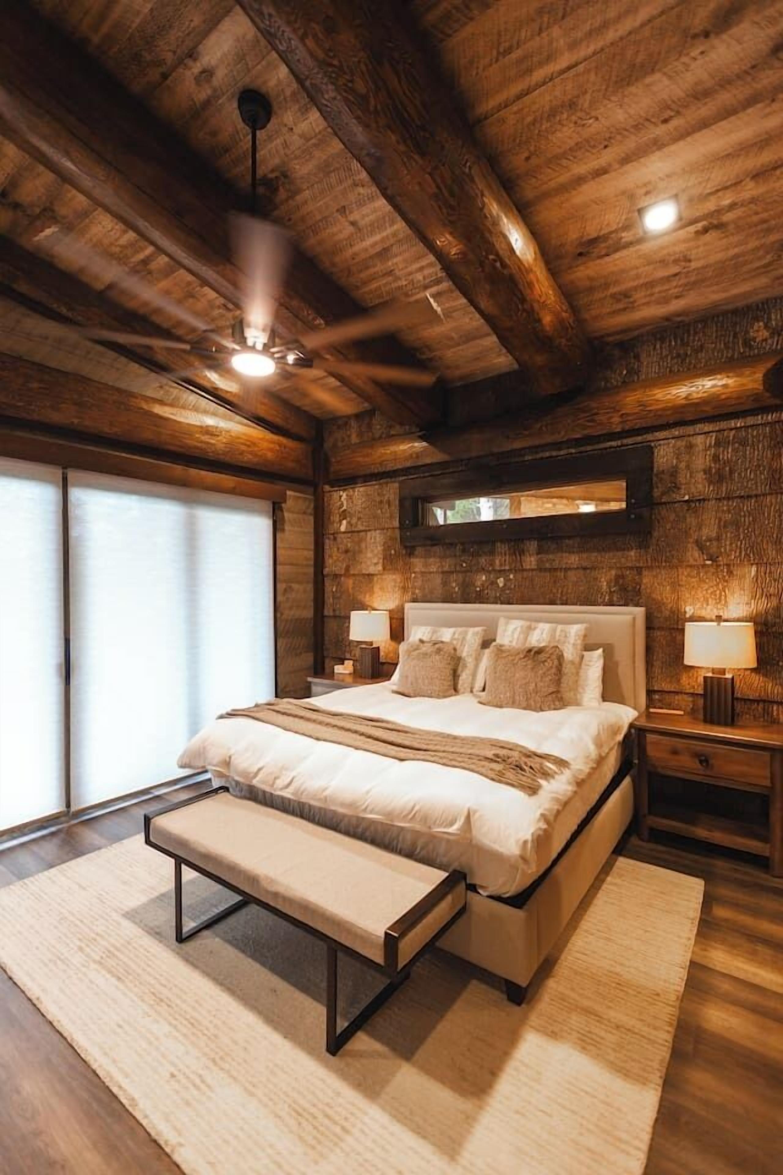 Un dormitorio con techo de madera, una cama con banco, lámparas de noche y una ventana con persianas.
