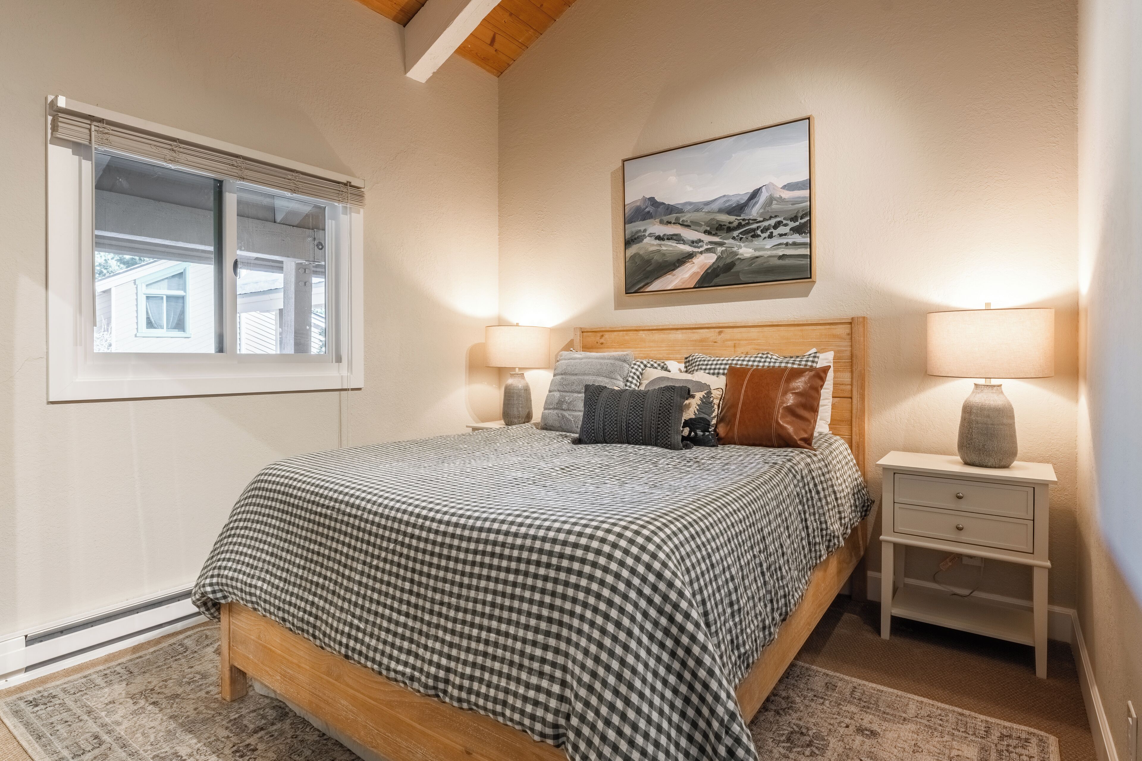Um quarto com cama de madeira, roupa de cama xadrez, duas luminárias de cabeceira, uma mesa de cabeceira, uma janela e uma pintura de paisagem na parede.