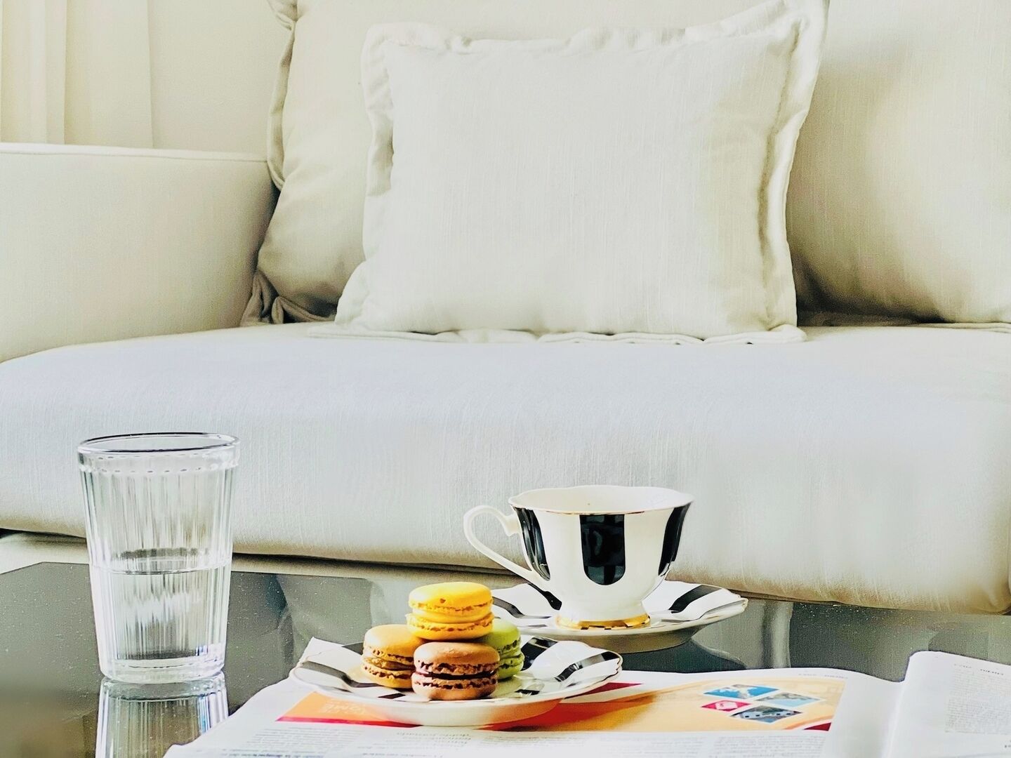 A neatly arranged breakfast setting with a glass of water, a cup and saucer, and a plate of macarons on a glass table.
