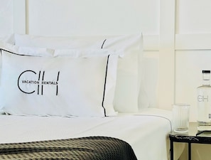 Quarto de hotel com cama, travesseiro com a inscrição 'Vacation Rentals', mesa de cabeceira com garrafa e copo, e manta de tricô preta.