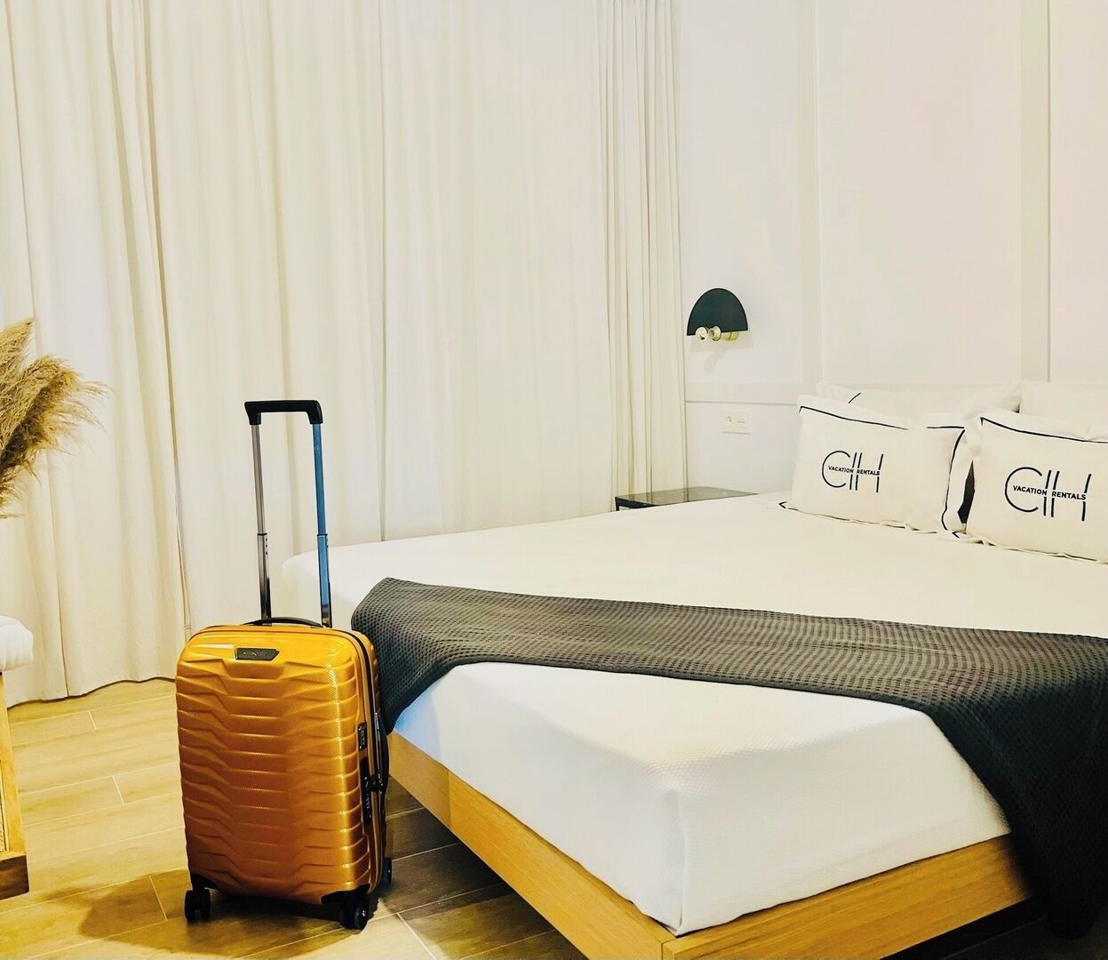 A neatly made bed with a white and gray blanket, a yellow suitcase, and pillows with "Vacation Rentals" text.