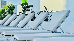 A row of white sun loungers with metal frames, positioned on a flat surface with potted plants in the background. - CIH Apartments (Puerta del Carmen)