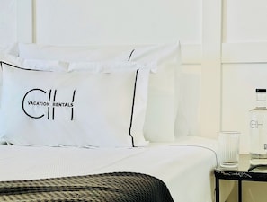 Un lit bien fait, avec du linge de lit blanc et des oreillers, dont l’un est brodé du mot « Locations de vacances ». On peut voir un verre d’eau et une bouteille sur une petite table placée à côté du lit.