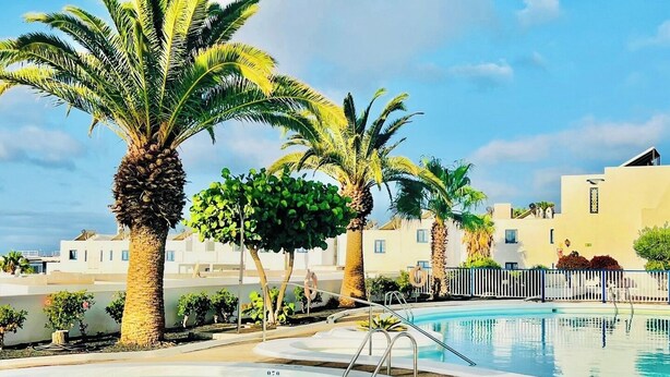 Zona de piscina con palmeras, un solárium y un edificio al fondo.