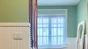 Una habitación con un armario de madera, una ventana con persianas, un pequeño banco y un espejo apoyado en la pared.