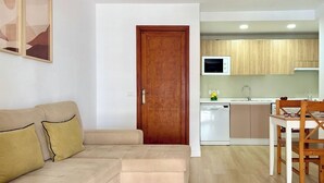 Una sala de estar con un sofá color beige, una puerta de madera y una zona de cocina con armarios de madera y un microondas blanco.