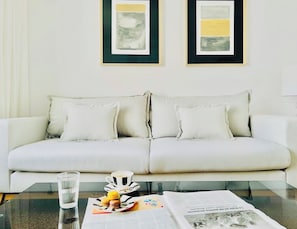 A white sofa with two pillows, a glass table with a cup and macarons, and two framed abstract paintings on the wall.