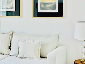 A white sofa with two pillows, a lamp on a wooden side table, and two framed abstract paintings on the wall.