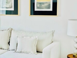 A white sofa with three pillows, a lamp on a wooden side table, and two framed abstract paintings on the wall.