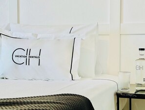 A hotel room with a bed, a pillow with "Vacation Rentals" text, a nightstand with a bottle and glass, and a black woven throw blanket.