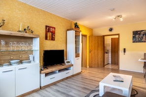 A modern living room with a TV, white cabinets, a wooden shelf with glasses, and a small table.