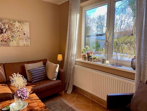 Ein gemütliches Wohnzimmer mit einem braunen Ledersofa, einem Glastisch mit einer Vase, einem Fenster mit Pflanzen und einer Heizung.