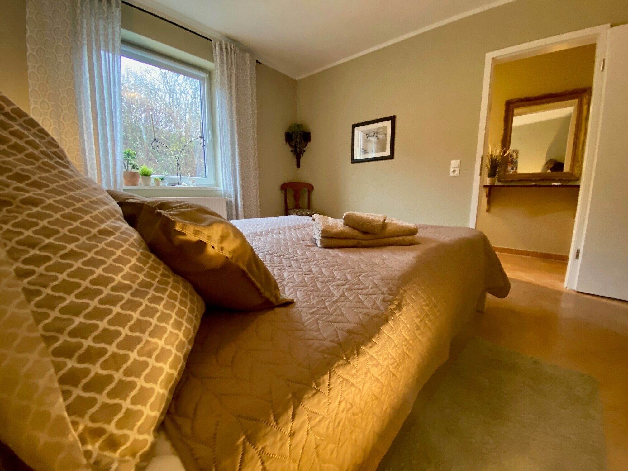 A bedroom with a large bed, a window with curtains, a framed picture on the wall, and a mirror in the adjacent room.