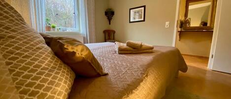A bedroom with a large bed, a window with curtains, a framed picture on the wall, and a mirror in the adjacent room.