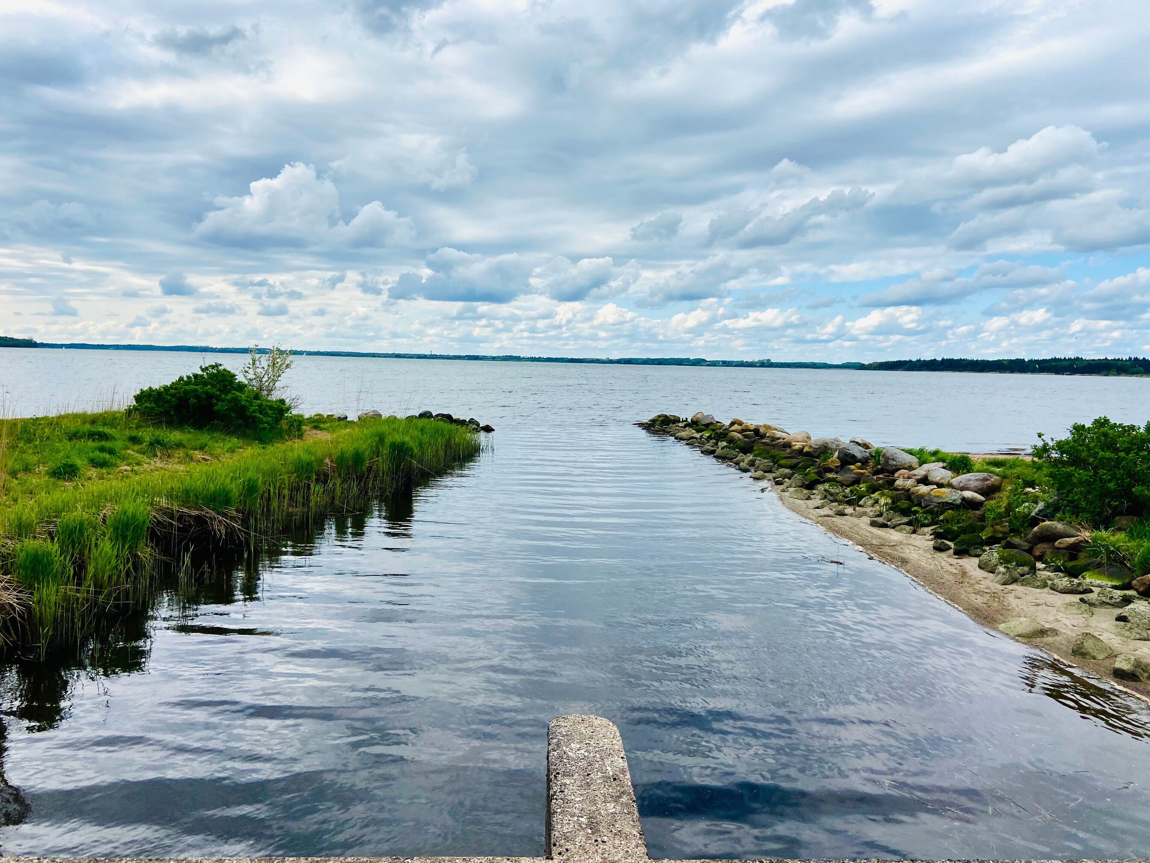 Stille vandvej med en betonpromenade, der fører til en bro, omgivet af græsarealer og sten.