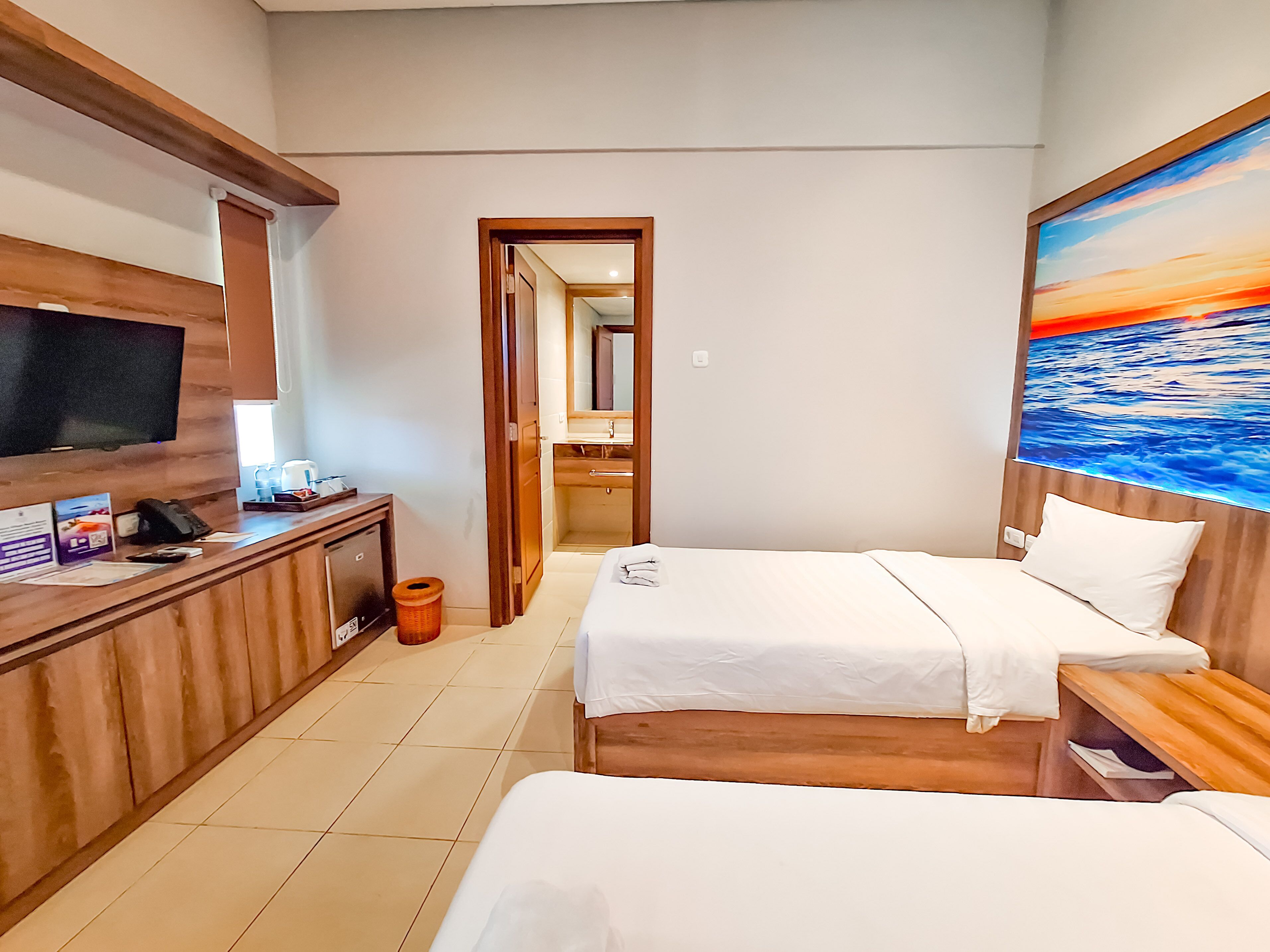A hotel room with two beds, a TV, a desk with a computer, and a painting of a sunset over the ocean.
