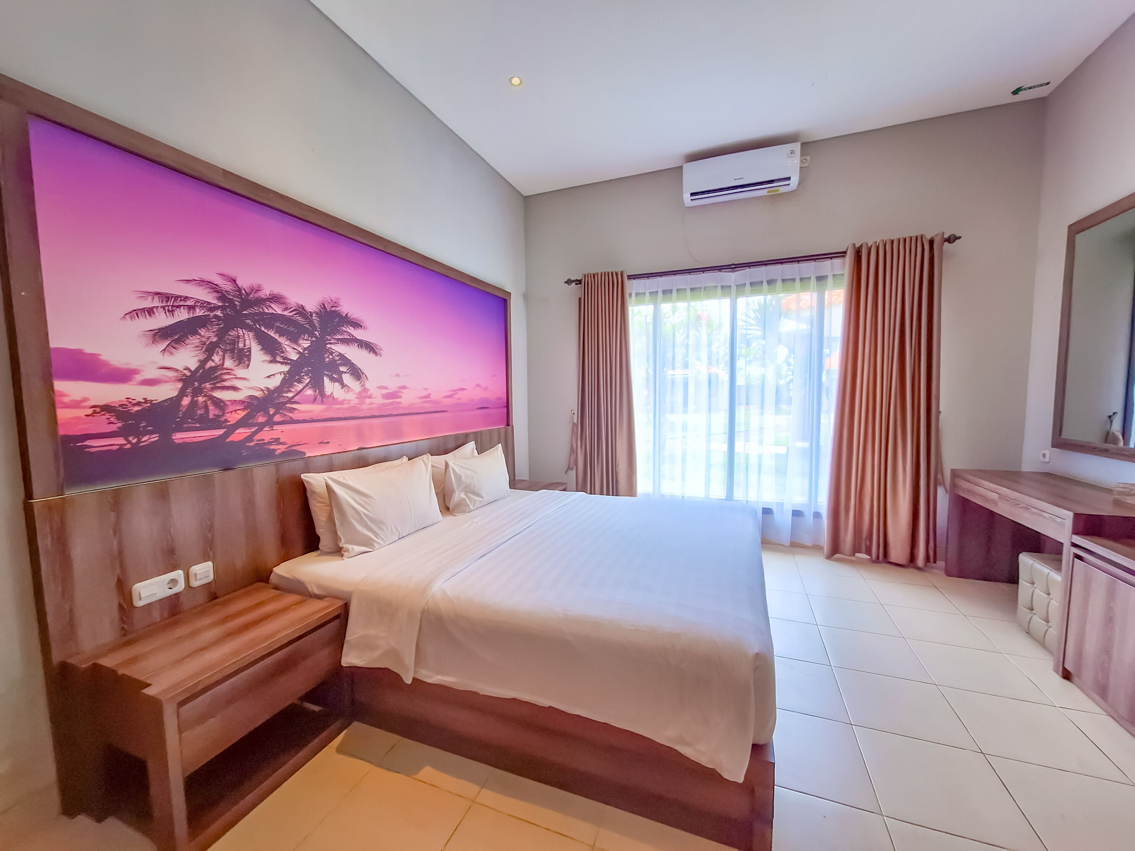 A hotel room with a large bed, a wooden bench, a desk with a chair, and a large window with curtains.
