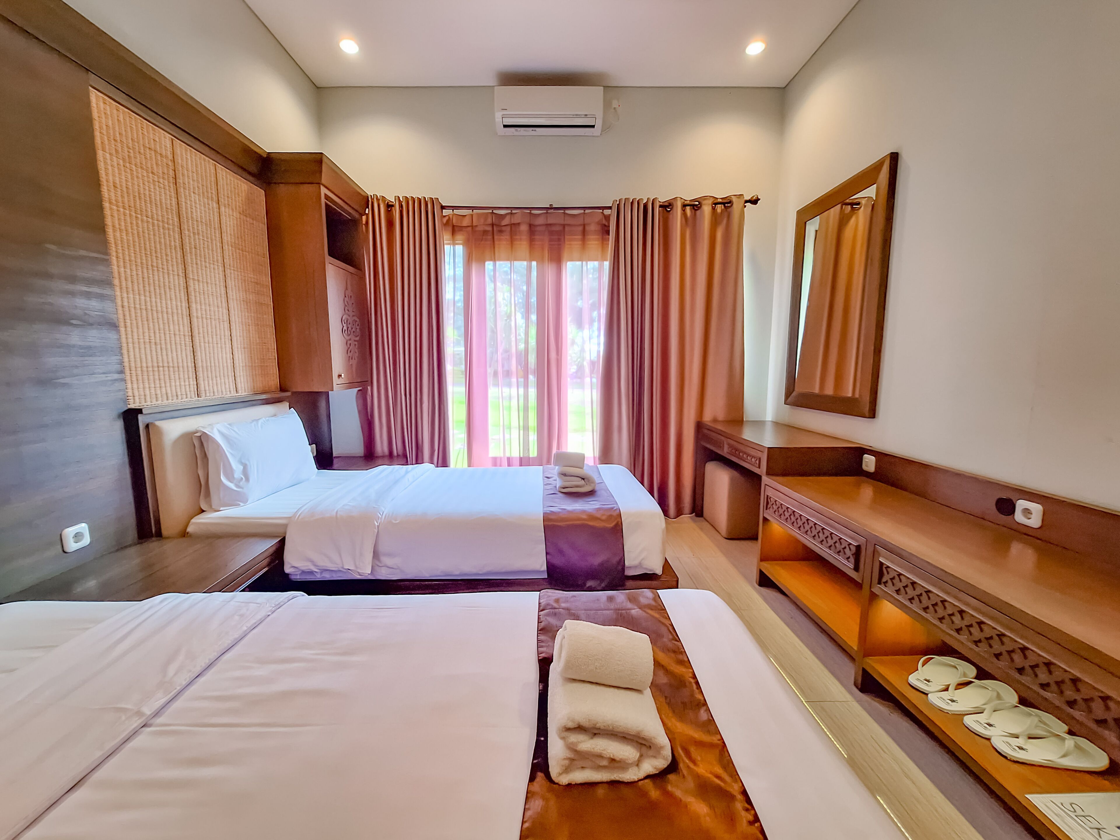 A hotel room with two beds, a wooden headboard, a large mirror, and a window with curtains.
