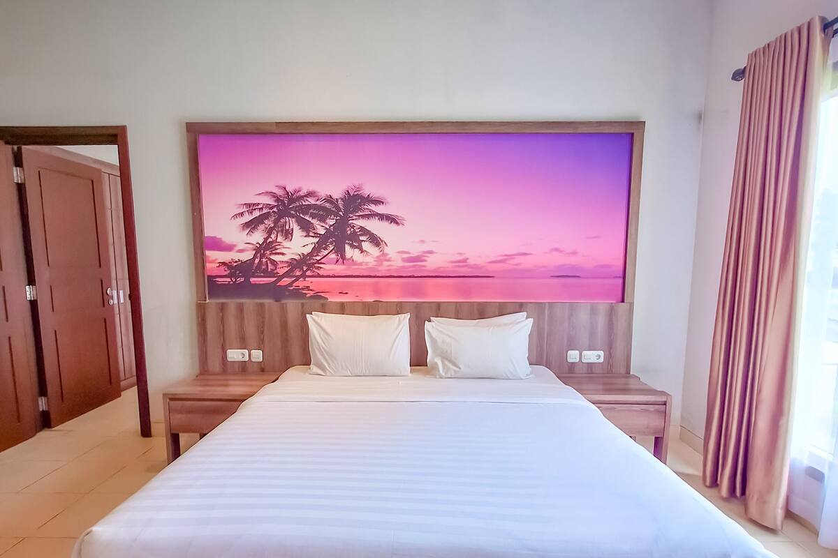 A large bed with a wooden frame, white bedding, and a headboard with a sunset painting. There are two bedside tables with lamps, a wooden nightstand, and a door to the left.