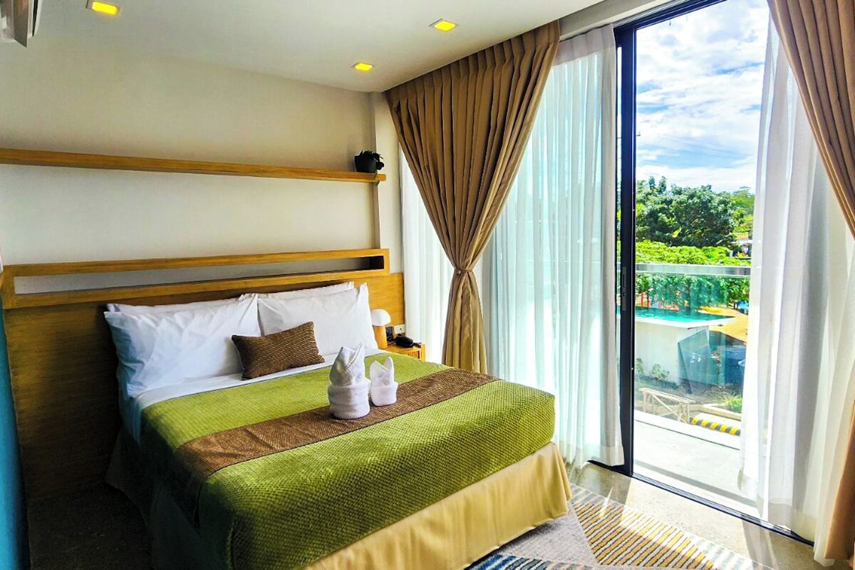 A hotel room with a bed, a nightstand, a window with curtains, and a view of greenery outside.