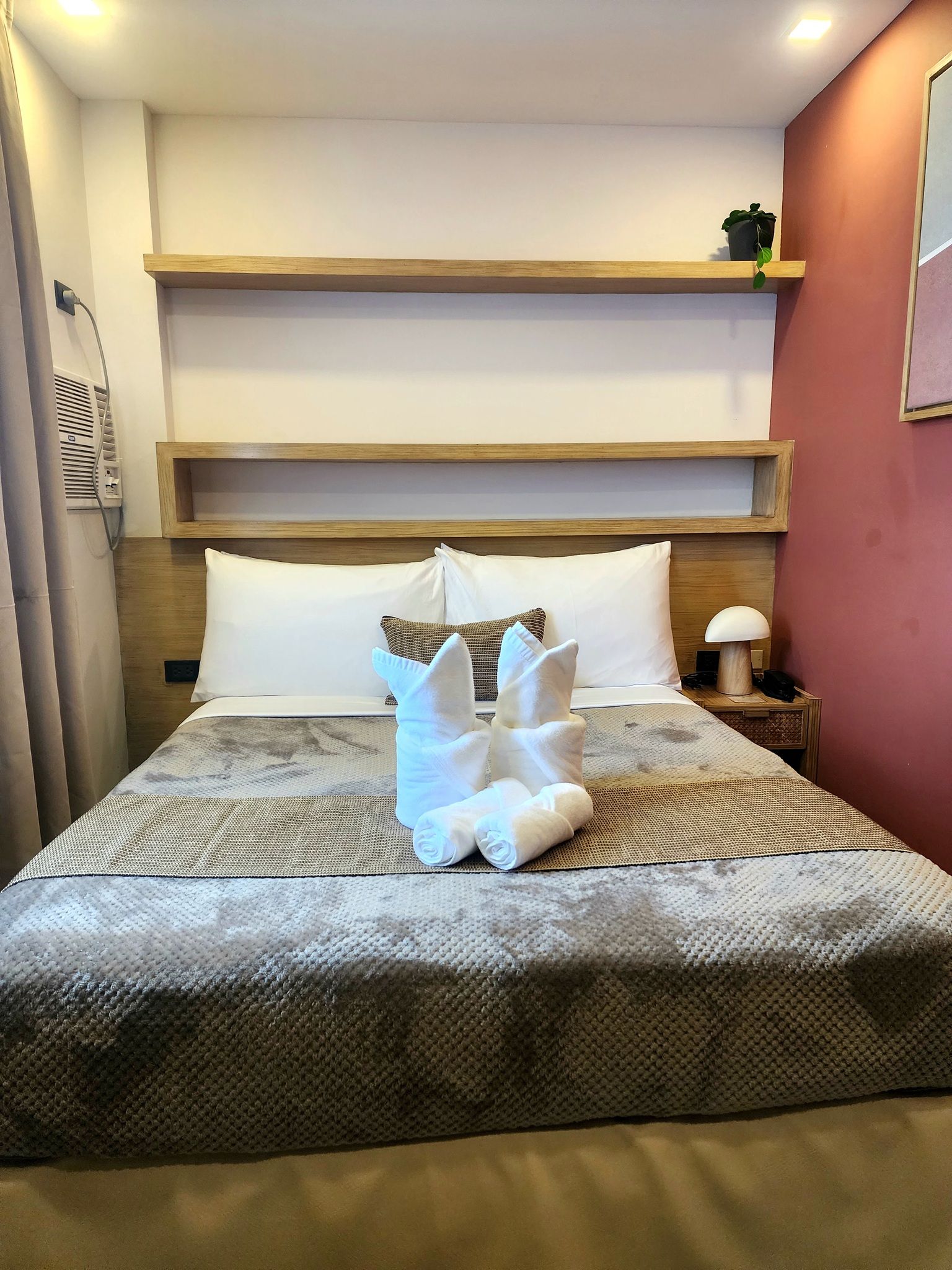 Quarto de hotel com cama, almofadas, abajur e prateleira fixada na parede.
