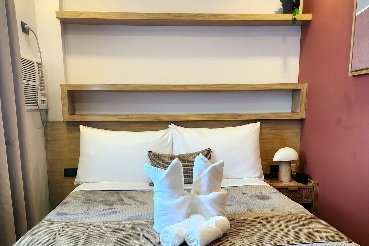 A hotel room with a bed, a nightstand, a lamp, and a wall-mounted shelf.