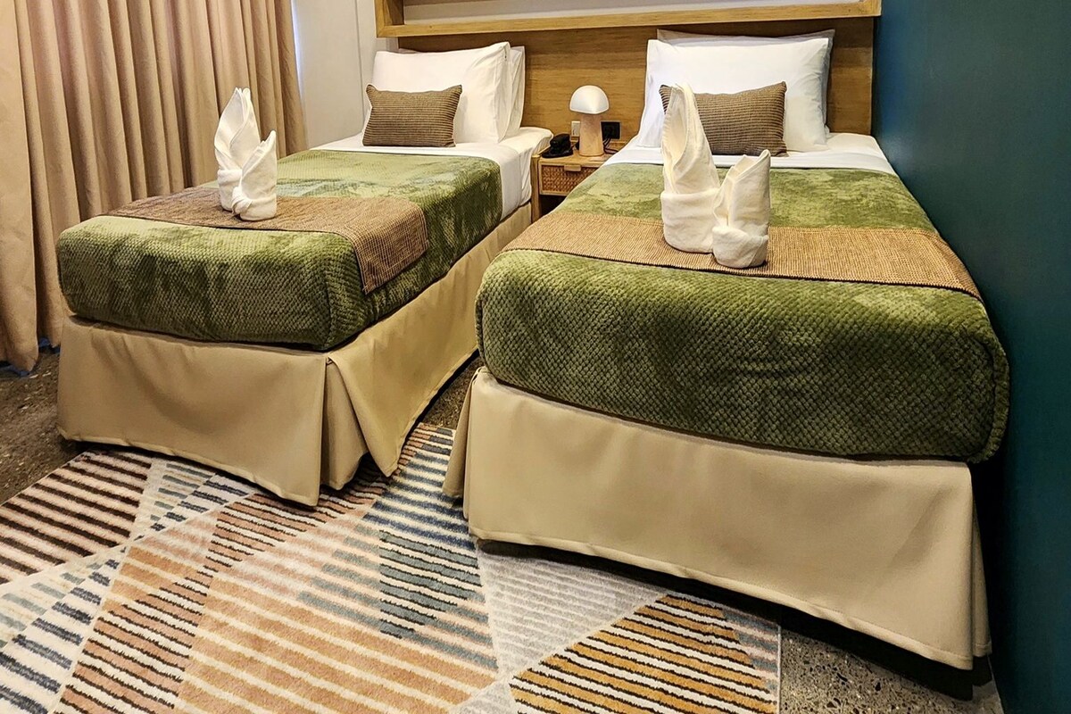 A hotel room with two beds, a nightstand, a lamp, and a window with curtains.
