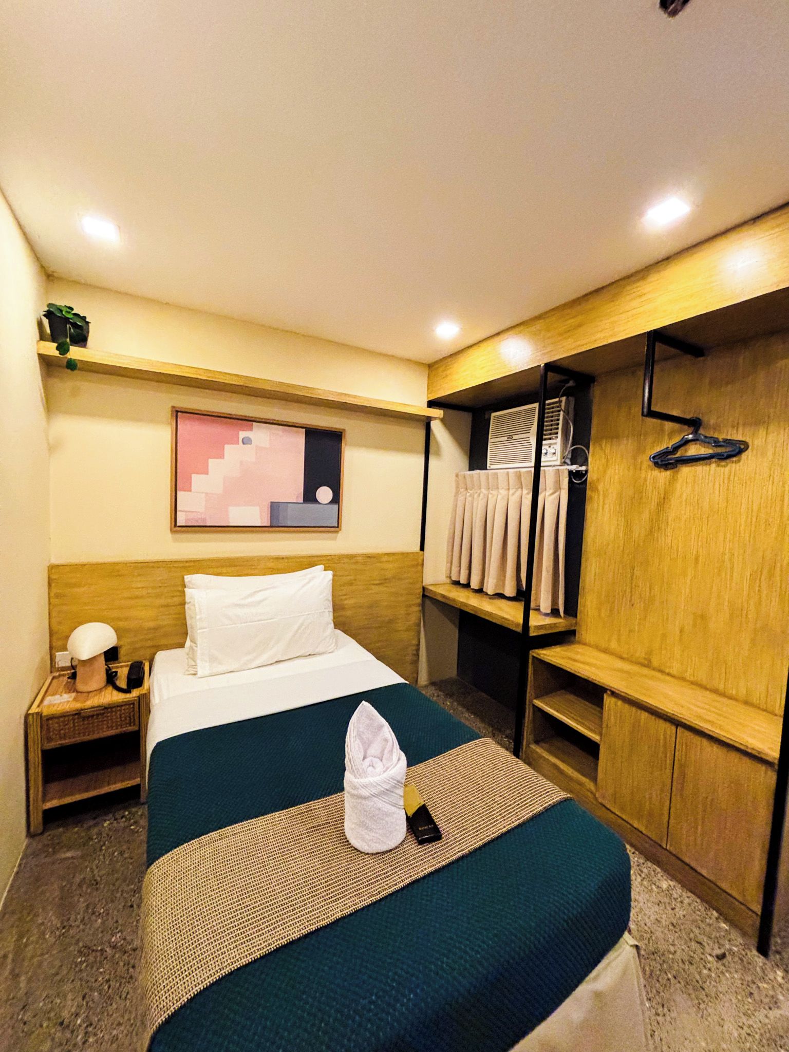 A hotel room with a bed, a nightstand, a wall-mounted shelf with a plant, a radiator, and a painting.