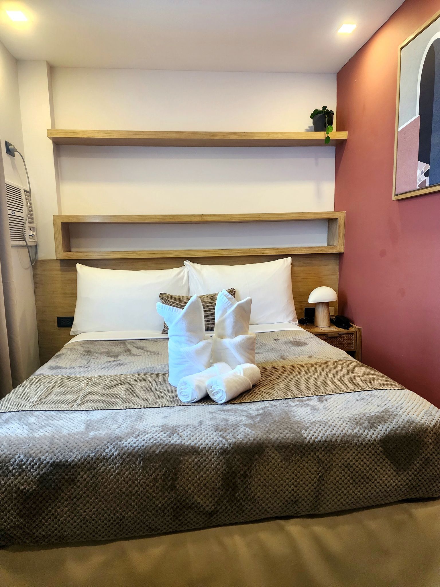 A hotel room with a bed, pillows, a nightstand, a lamp, and a wall-mounted shelf.