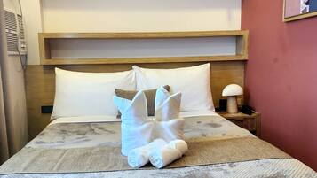 A hotel room with a bed, pillows, a nightstand, a lamp, and a wall-mounted shelf.