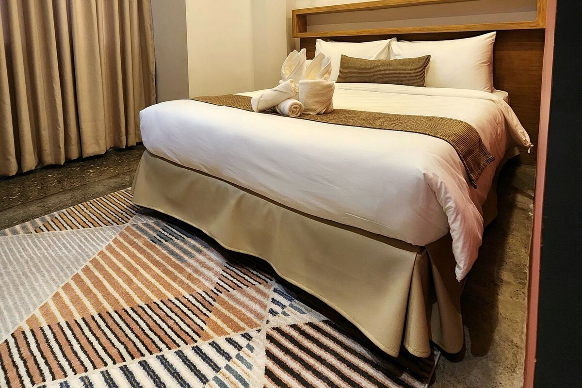 A hotel room with a bed, a bedside table, a wall-mounted shelf, and a large window with curtains.