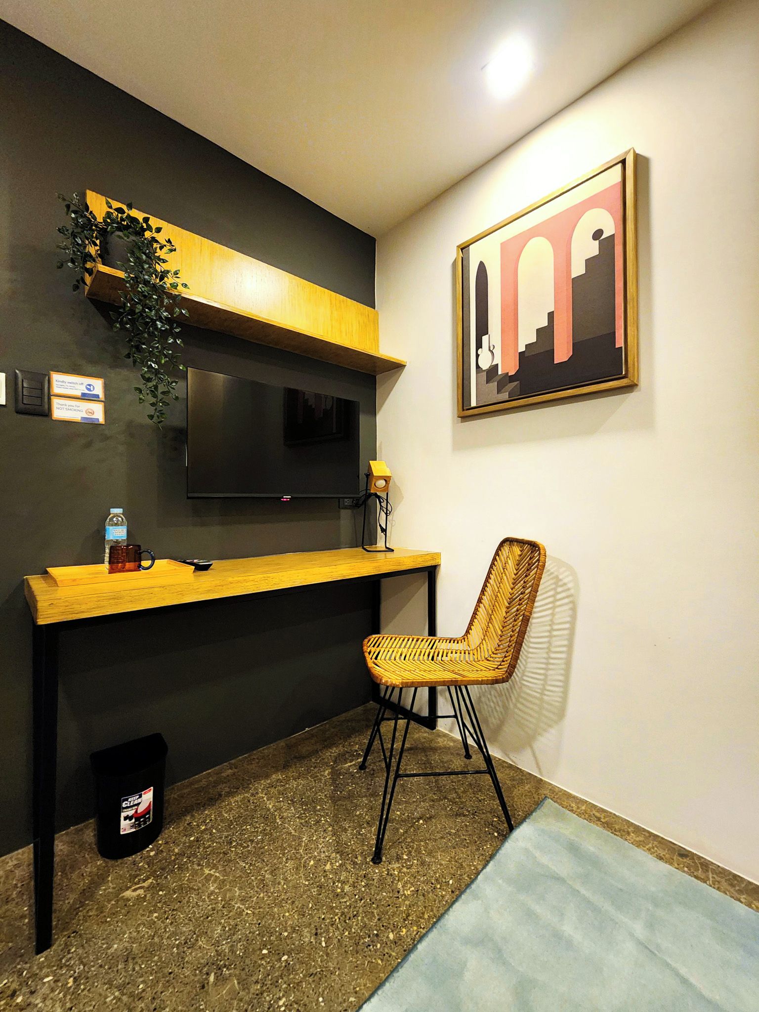 A modern room with a wooden desk, a television mounted on the wall, a wicker chair, and a framed artwork.