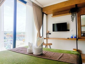 Une chambre d’hôtel dotée d’une grande fenêtre, d’un lit recouvert d’un tapis vert et marron, d’un téléviseur à écran plat fixé au mur et d’une petite étagère sur laquelle sont posés divers objets.
