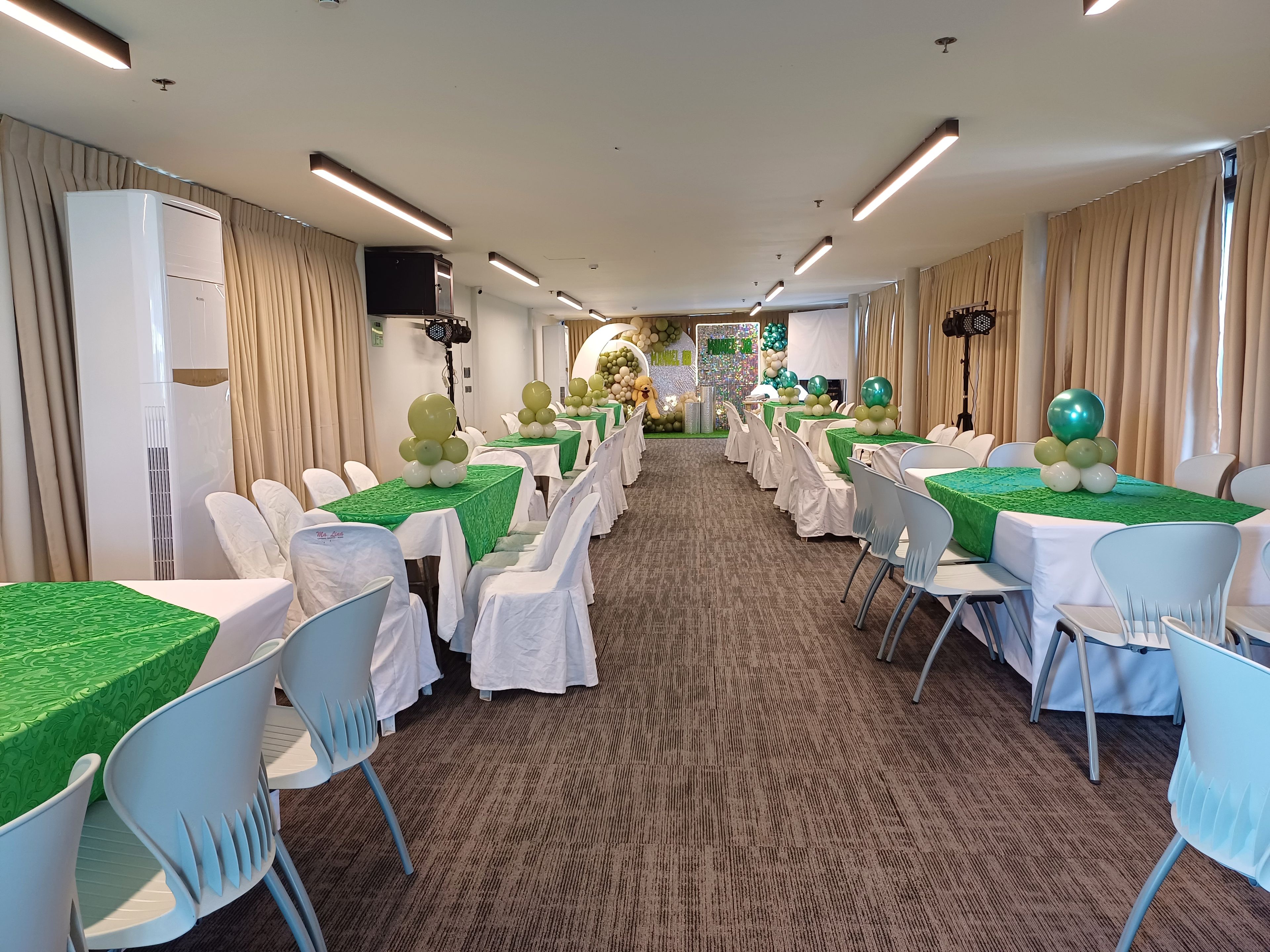 Salão de festas com mesas montadas para um evento formal, com piso carpete e disposição de mesas alongada.