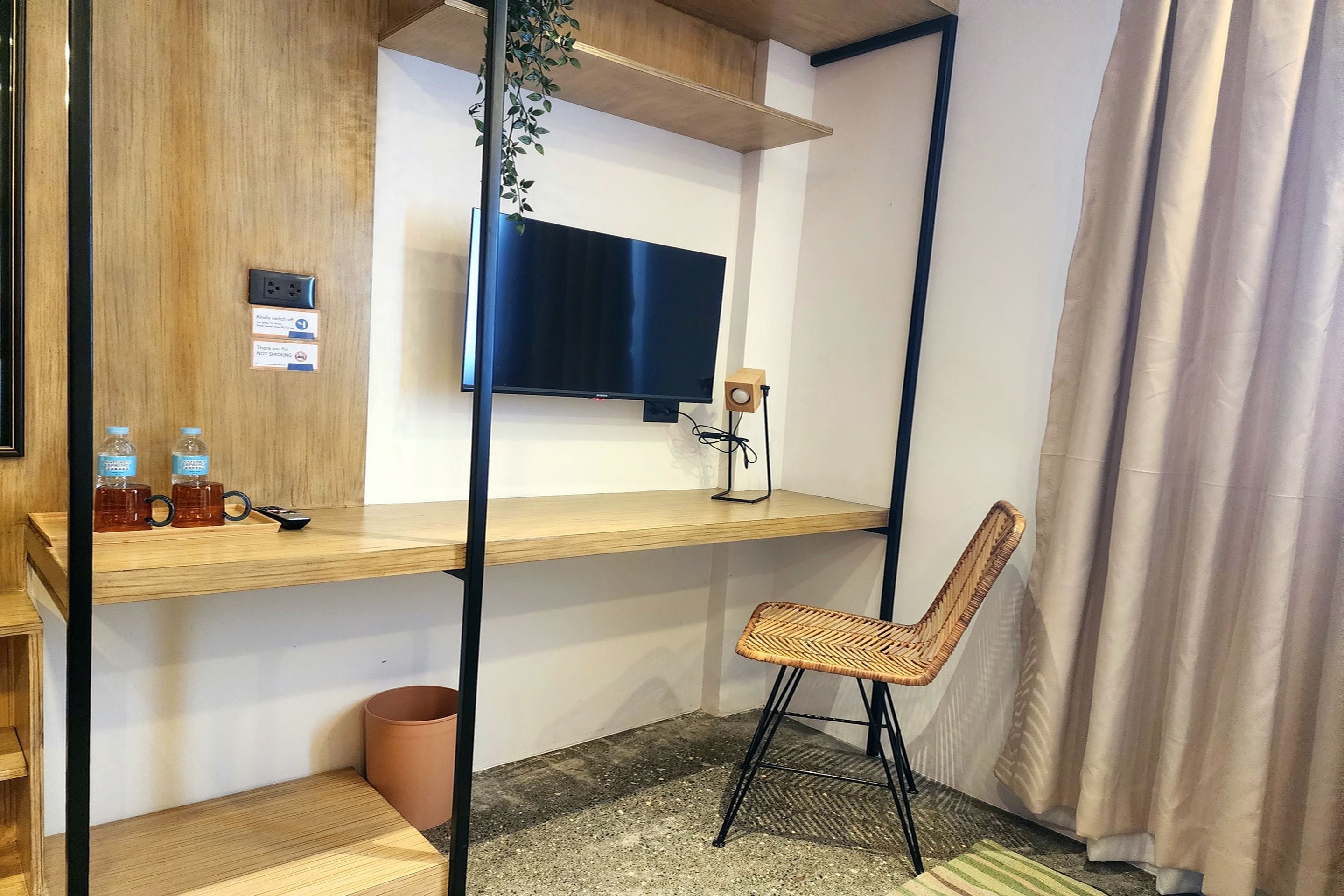 A compact hotel room with a wooden desk, a television mounted on the wall, a wicker chair, and a small trash bin.