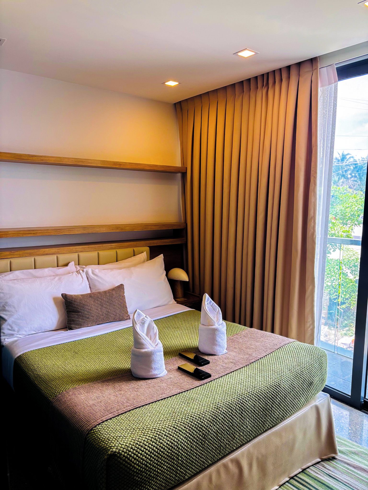 A hotel room with a large bed, a headboard, a window with curtains, and a view of greenery outside.
