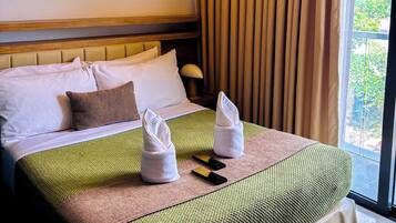 A hotel room with a large bed, a headboard, a window with curtains, and a view of greenery outside.