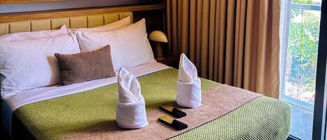 A hotel room with a large bed, a headboard, a window with curtains, and a view of greenery outside.