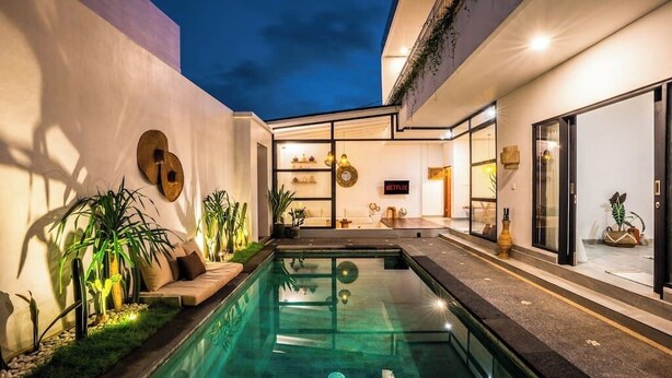 Un espace piscine extérieur moderne, doté d’une structure en verre, entouré de plantes et agrémenté d’un coin salon.