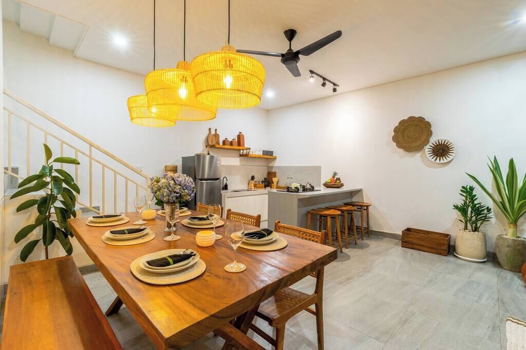Un coin repas avec une table en bois dressée pour quatre personnes, une cuisine en arrière-plan et un ventilateur de plafond.