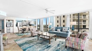A spacious living room with a sofa, armchairs, and a coffee table. There is a dining area with a table and chairs. The room has large windows and a view of the city.