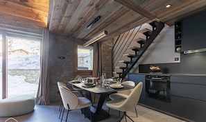 Una cocina moderna con encimera oscura y horno empotrado. Se ve un área de comedor con una mesa y sillas. Una escalera con barandilla negra conduce a un nivel superior.