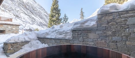 Un jacuzzi de paredes de piedra con un muelle de madera, rodeado de nieve y montañas.