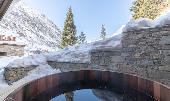 Un jacuzzi de paredes de piedra con un muelle de madera, rodeado de nieve y montañas.