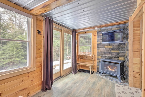 A cozy cabin interior with a stone fireplace, wooden walls, and large windows offering a forest view.