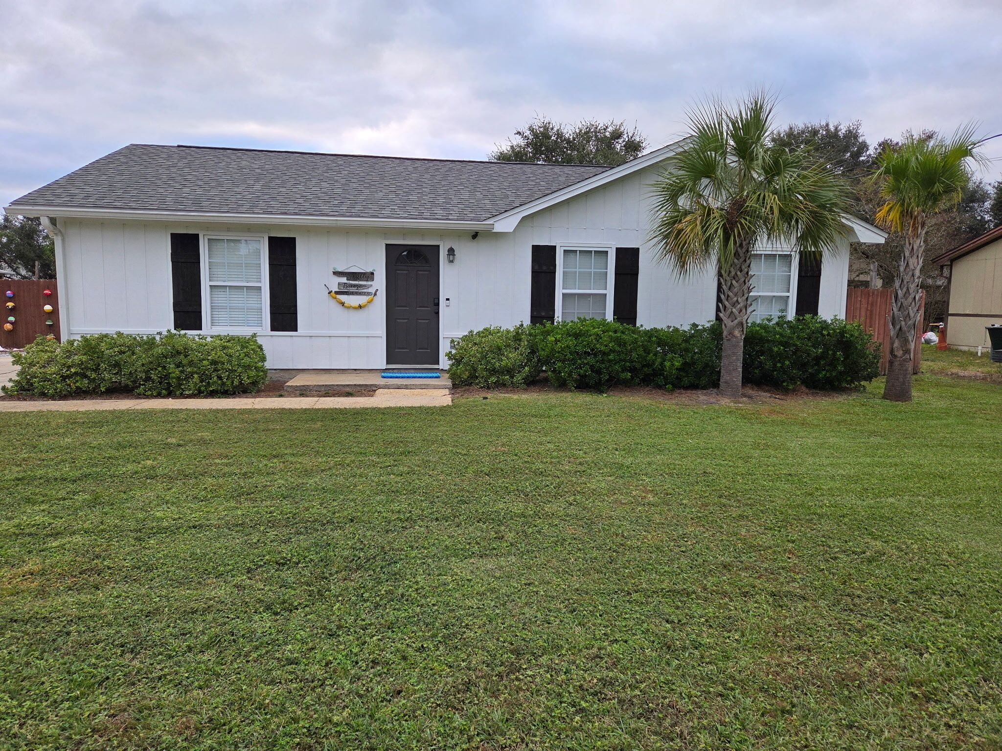 A single-story house with a dark roof, white siding, and black shutters. There are two small bushes in front of the house and a palm tree to the right.