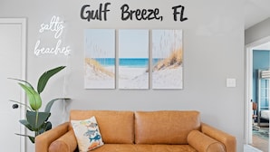 A tan leather sofa with a decorative pillow, a large green plant, and three beach-themed wall art pieces above it.