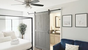 A bedroom with a four-poster bed, a ceiling fan, a sliding barn-style door, a blue sofa, and framed artwork on the wall.