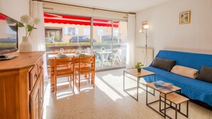 Un salón con sofá azul, mesa de comedor de madera y sillas, televisor en mueble de madera y balcón con toldo rojo.